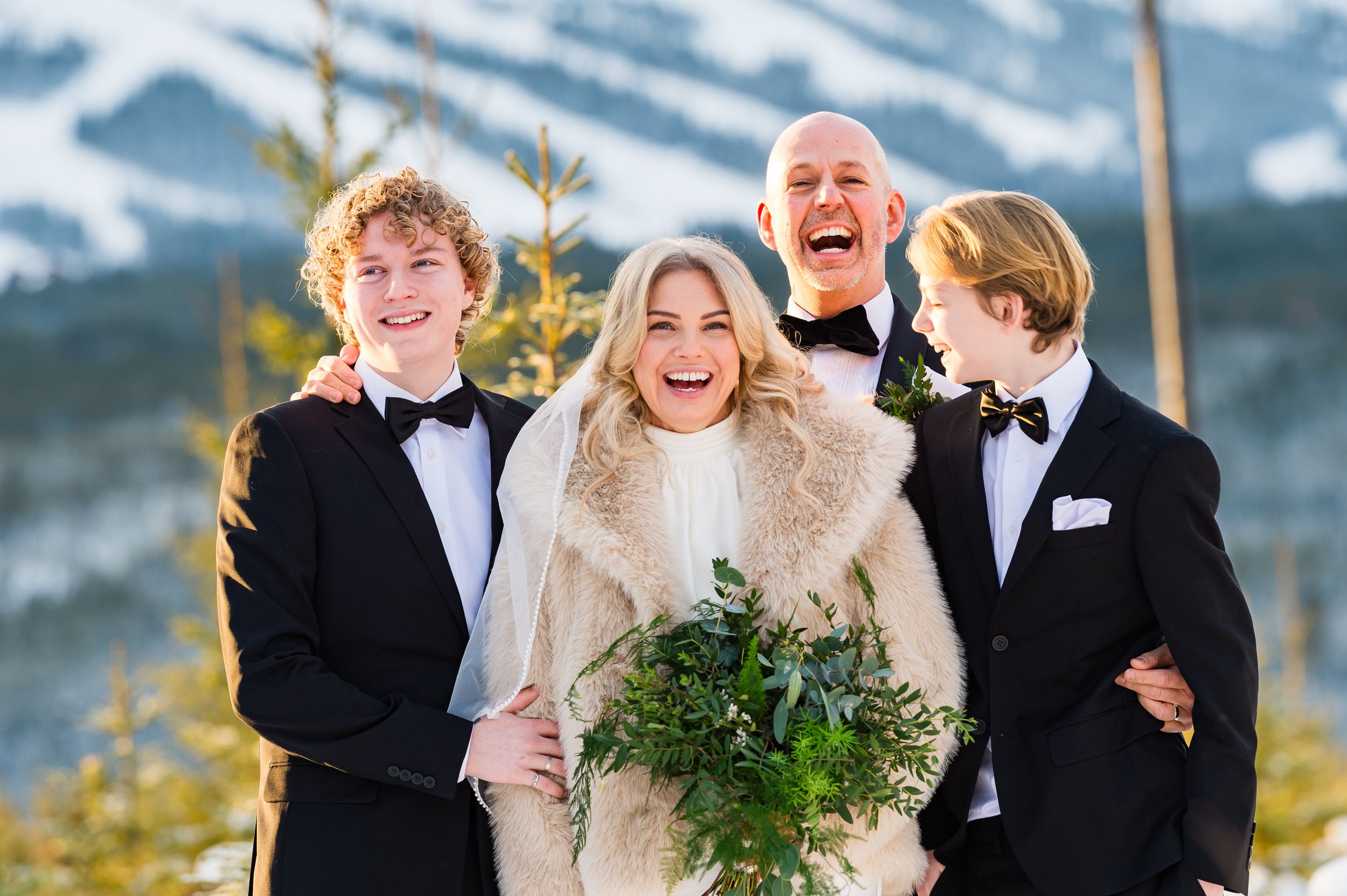 A joyful wedding party laughs together against a snowy mountain backdrop while holding a winter greenery bouquet.
