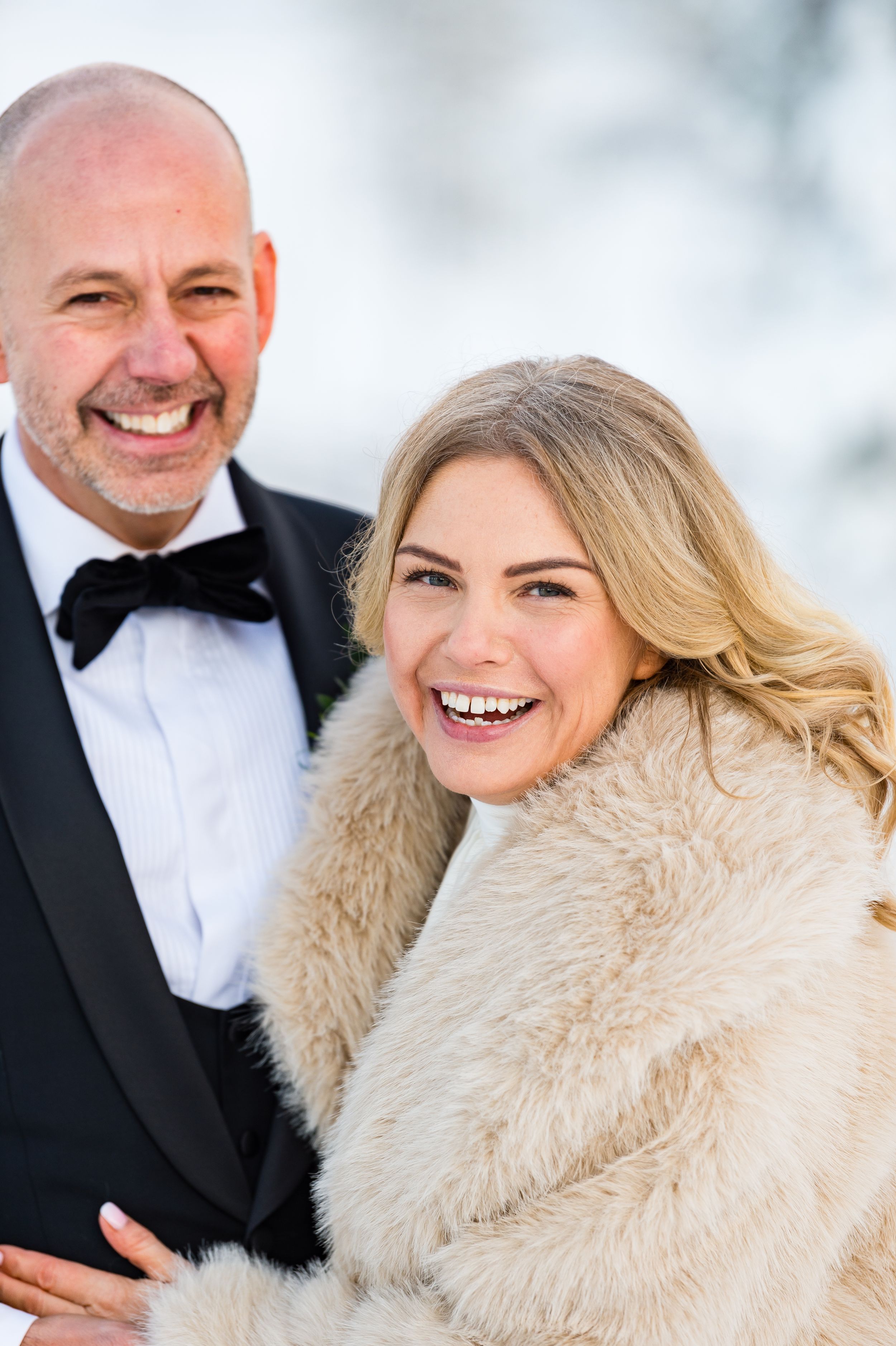 A couple shares joyful moments in formal attire with a white fur wrap against a snowy winter backdrop.