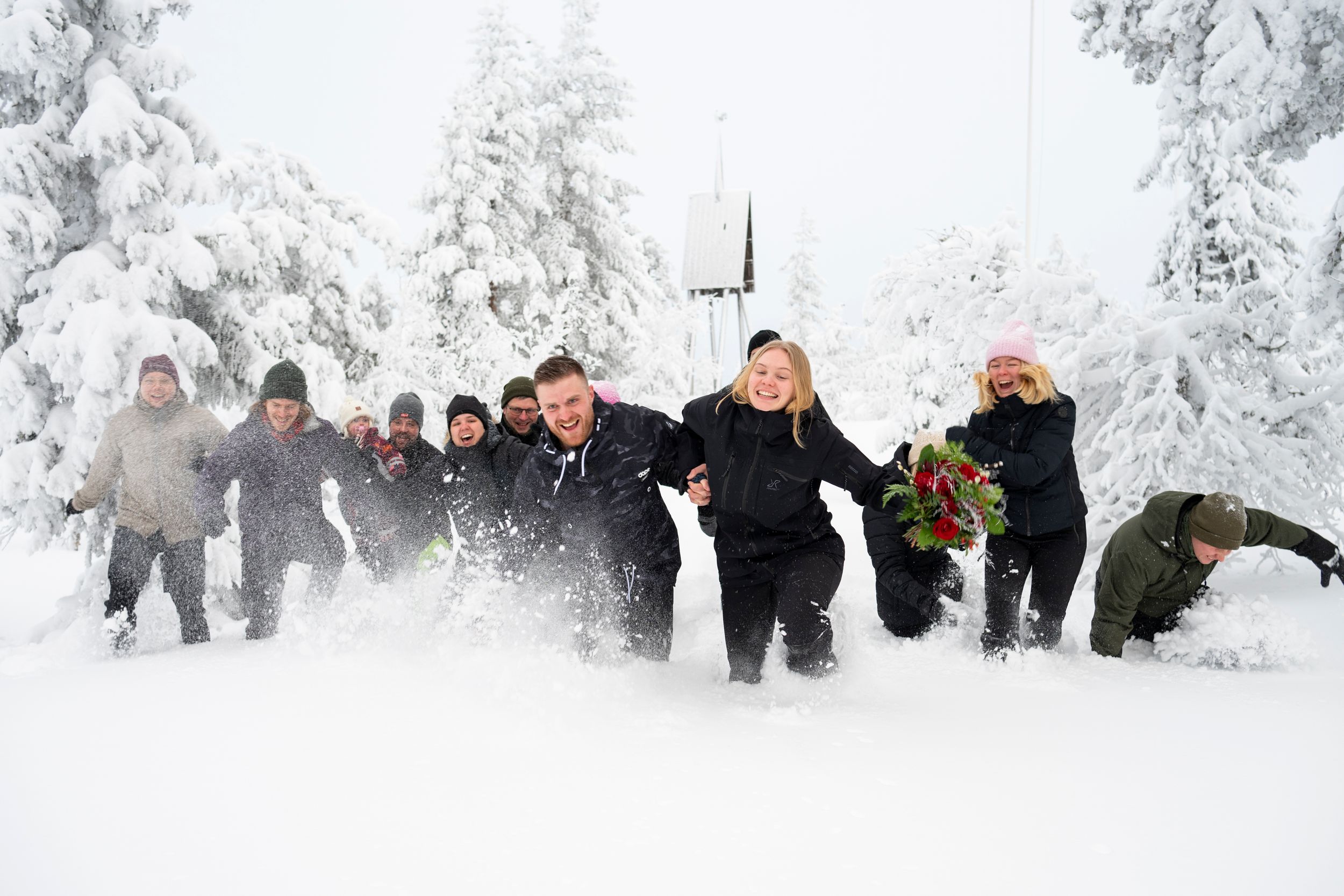 A group of wedding guests plays in deep snow surrounded by frost-covered trees in a winter landscape.