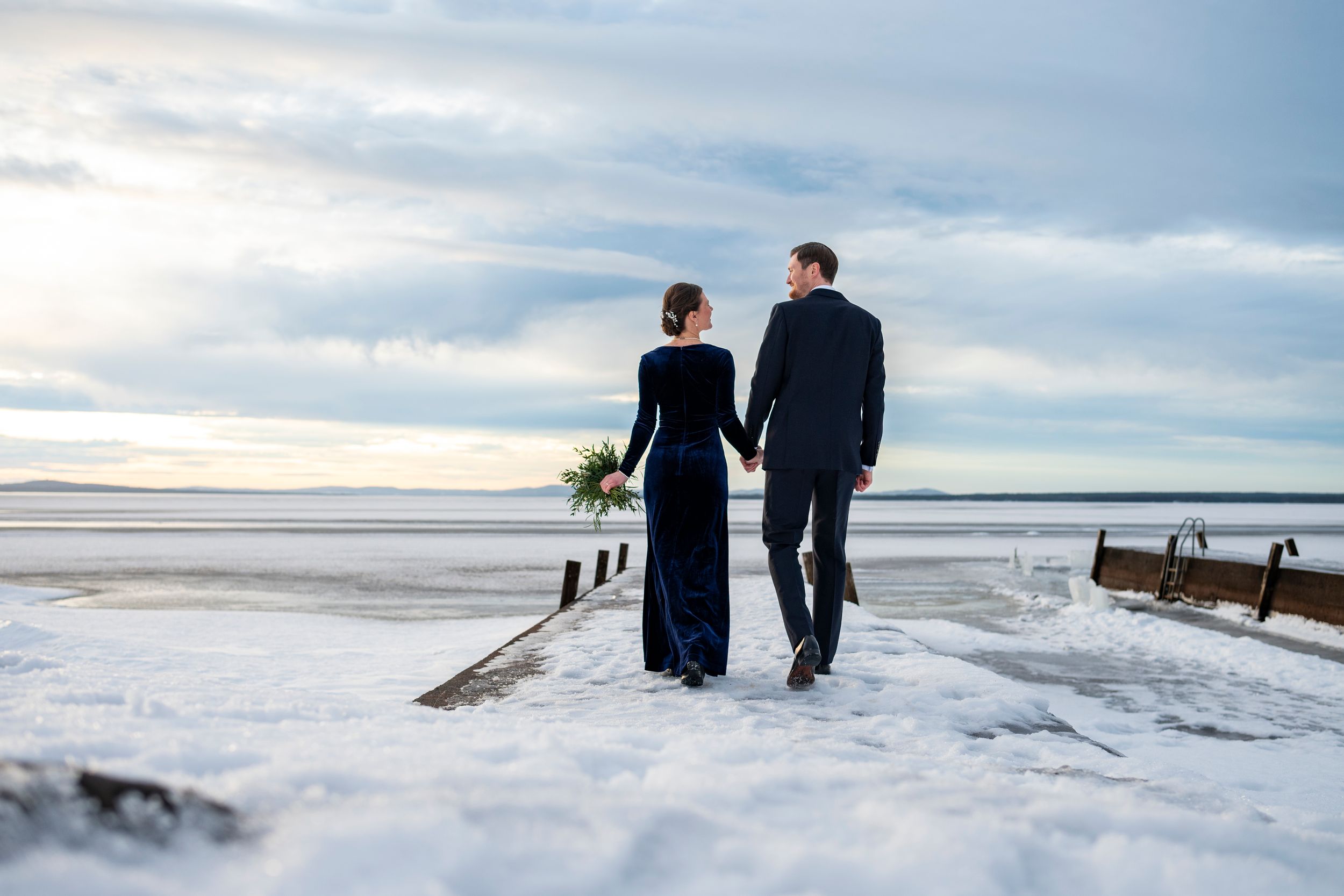 Two people walk along a snowy beach with a cloudy winter sky and ocean in the background.