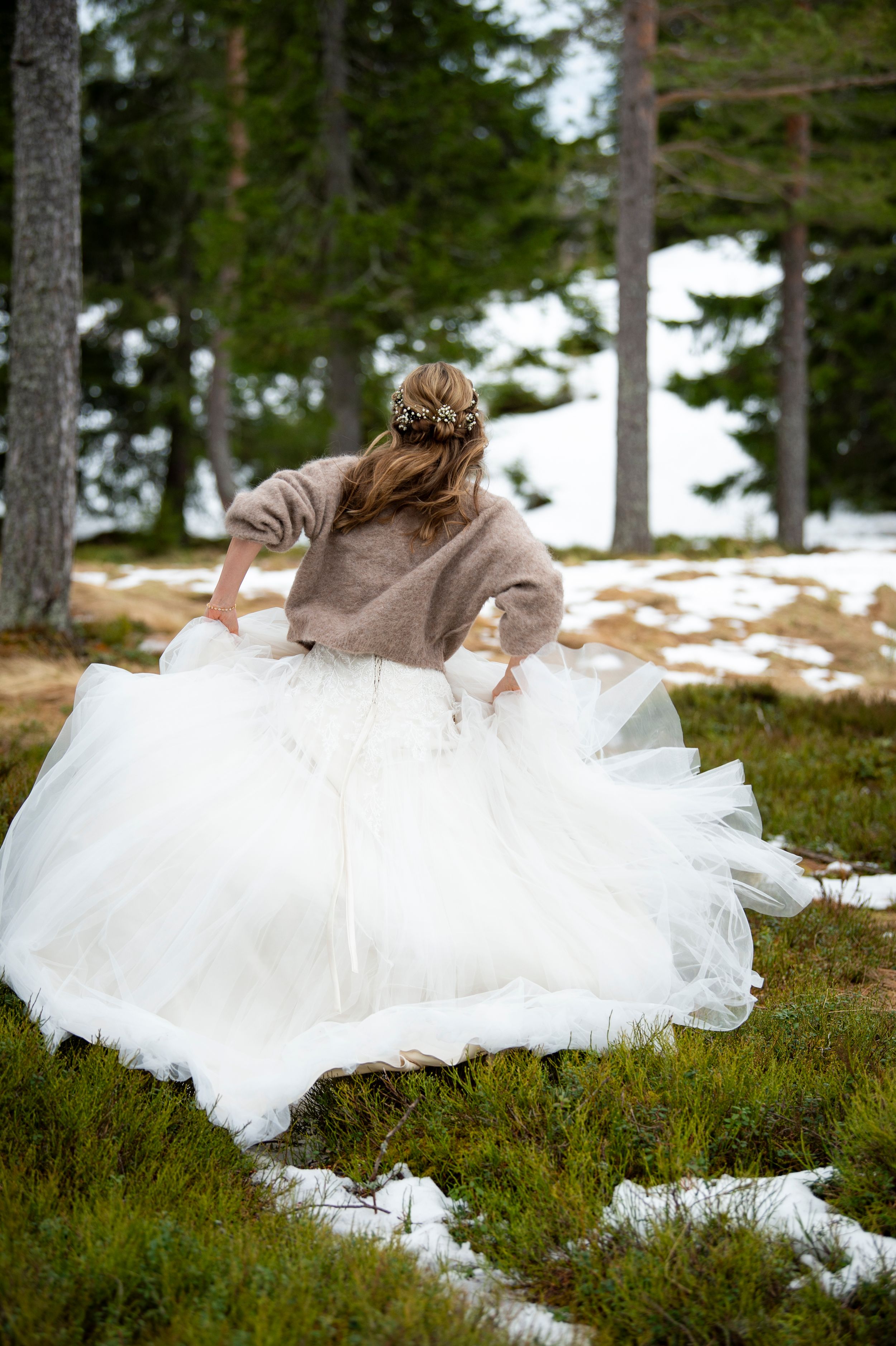 A white wedding dress flows across mossy ground in a snowy forest setting with pine trees in the background.