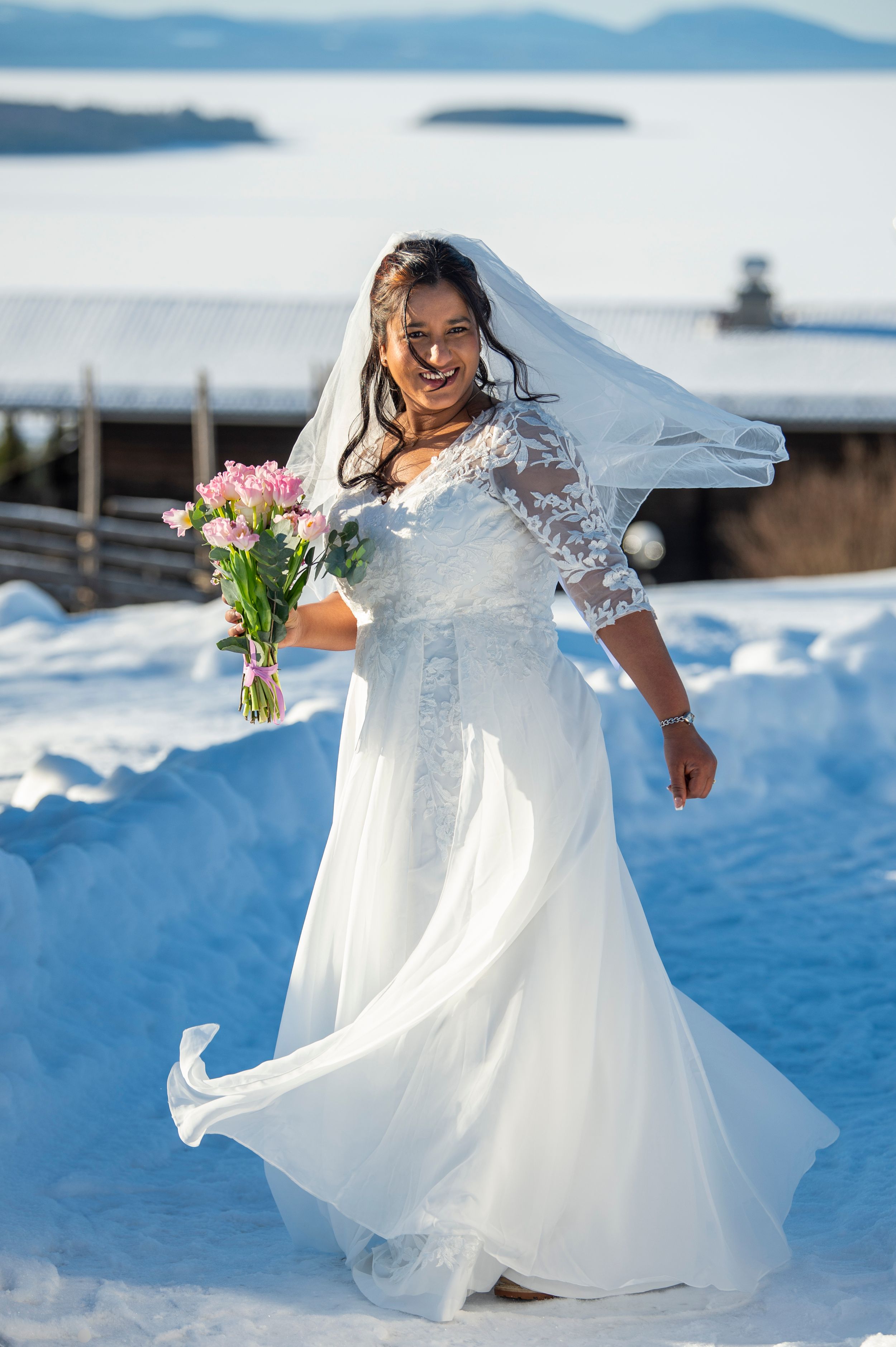 A flowing white wedding dress twirls gracefully in the snow with mountains and a lighthouse visible in the background.