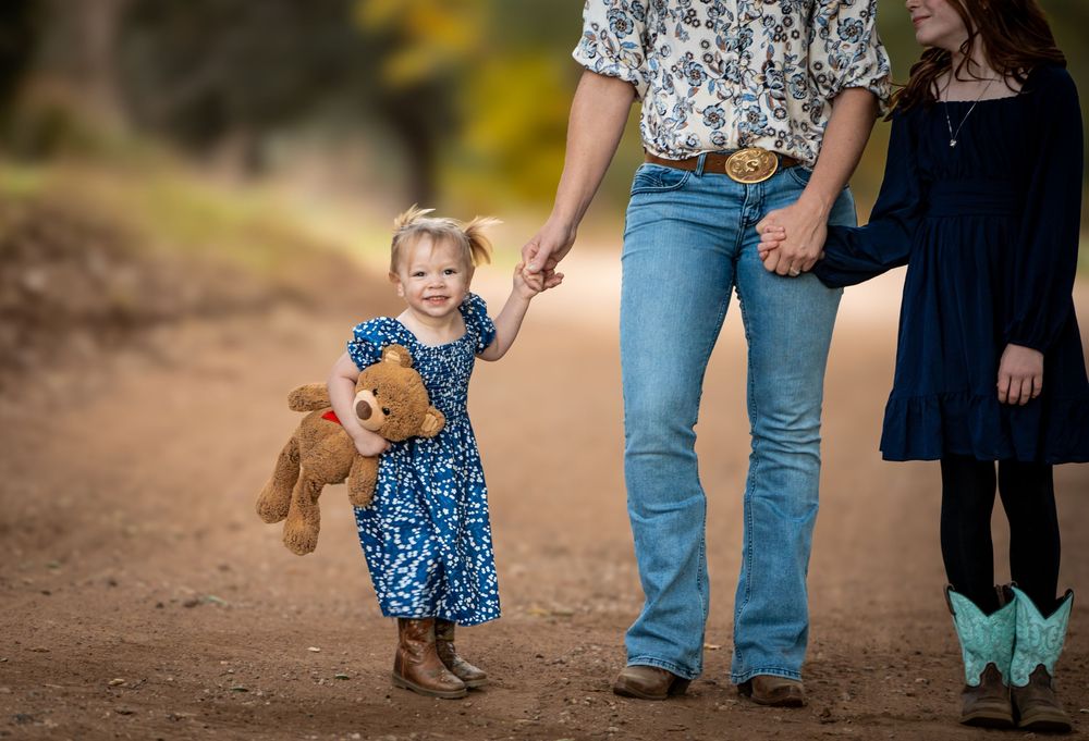 A toddler girl in a blue dress holding a teddy bear walks hand-in-hand with parents down a dirt path. family photos, Ramona CA