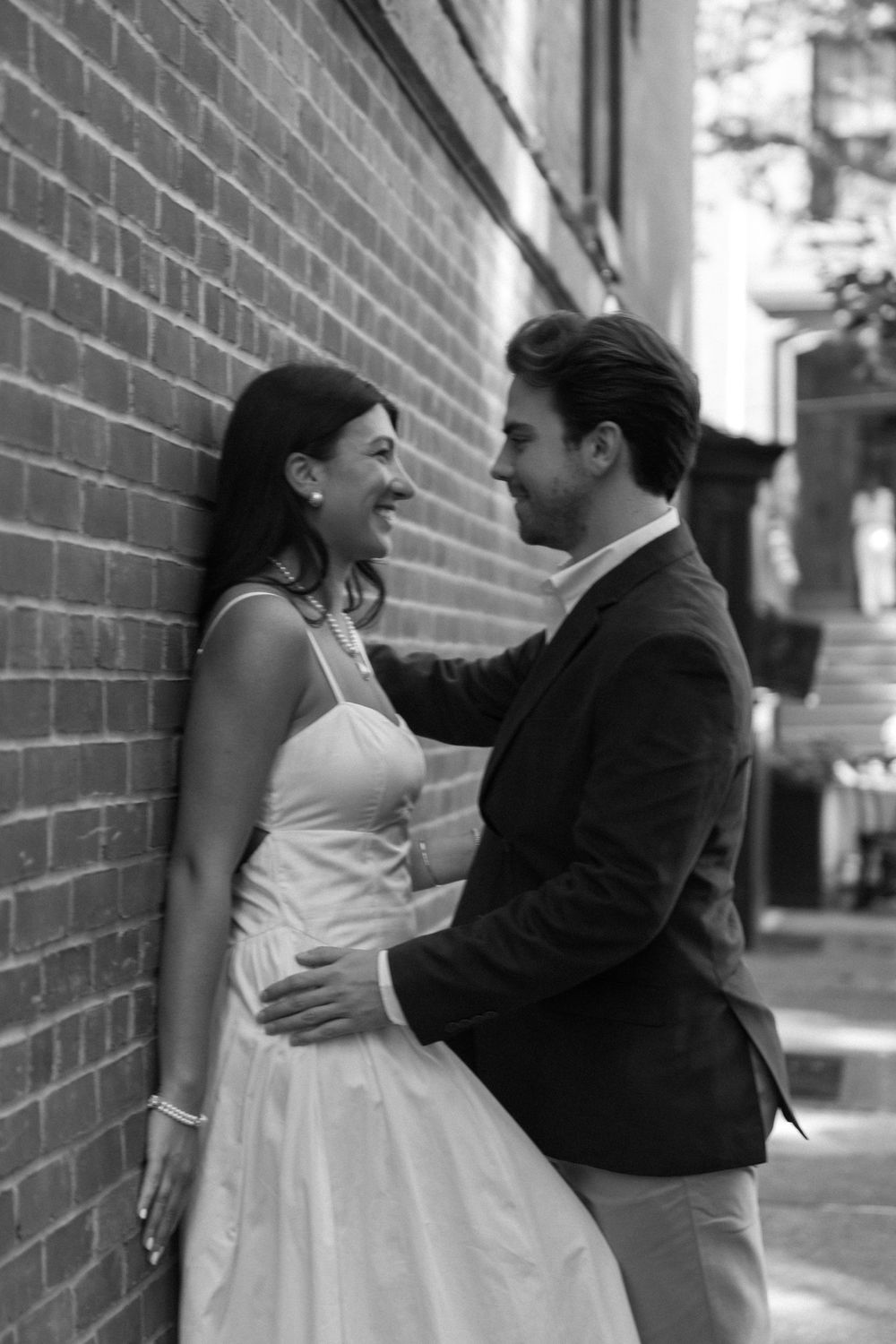 A couple shares a joyful moment against a brick wall, with her in a white dress and him in a dark blazer.
