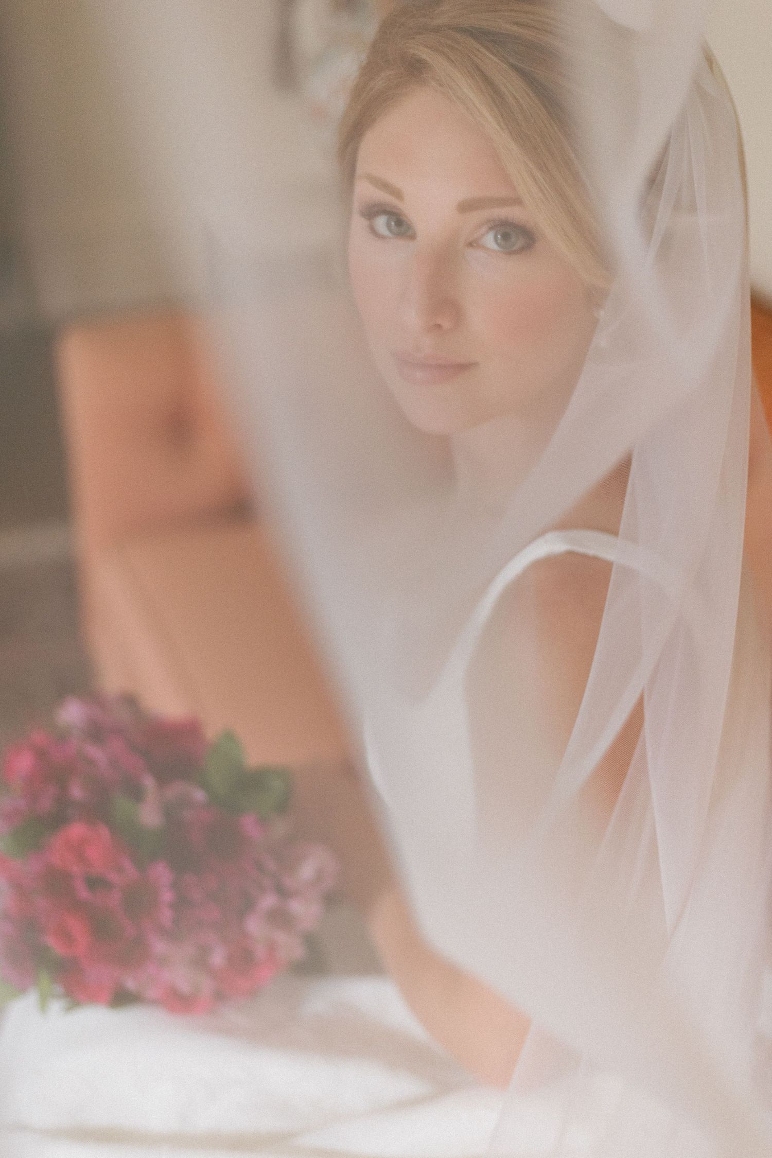 Ethereal bride gazes through soft white veil holding vibrant pink floral bouquet in dreamy portrait.