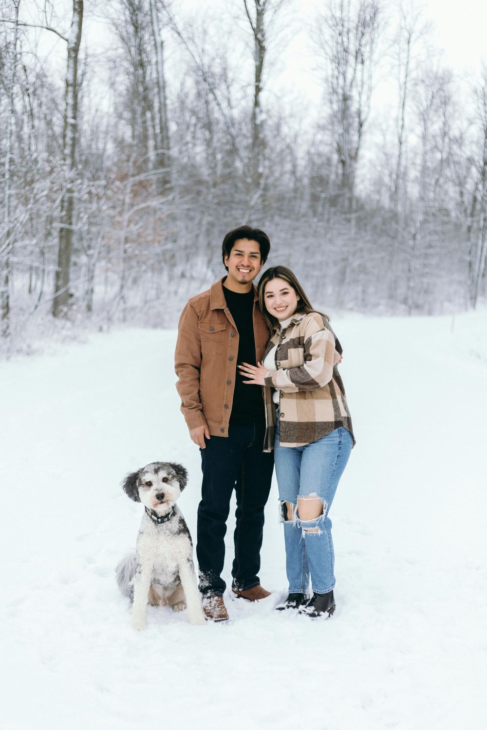 A couple and their scruffy terrier dog pose together in a snowy winter forest scene.