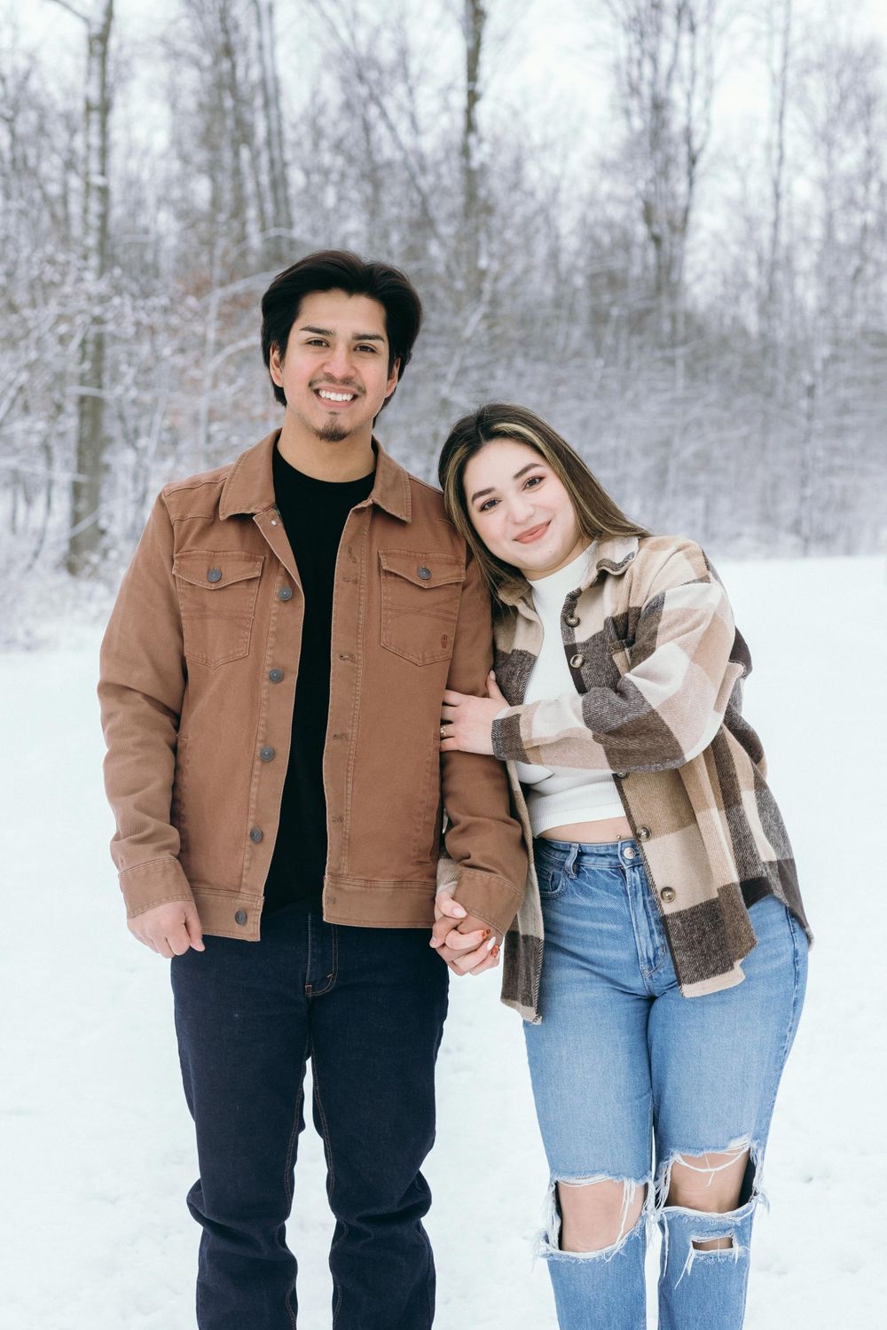 A couple stands together in a snowy winter forest scene wearing casual jackets and jeans.