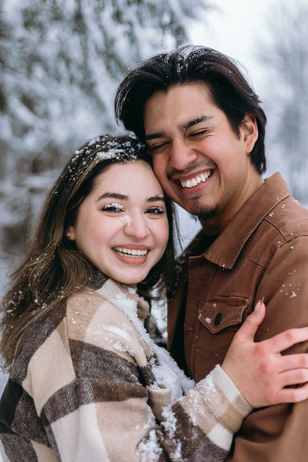 Happy couple laughing together in snow wearing warm sweaters and jackets during winter photo session.