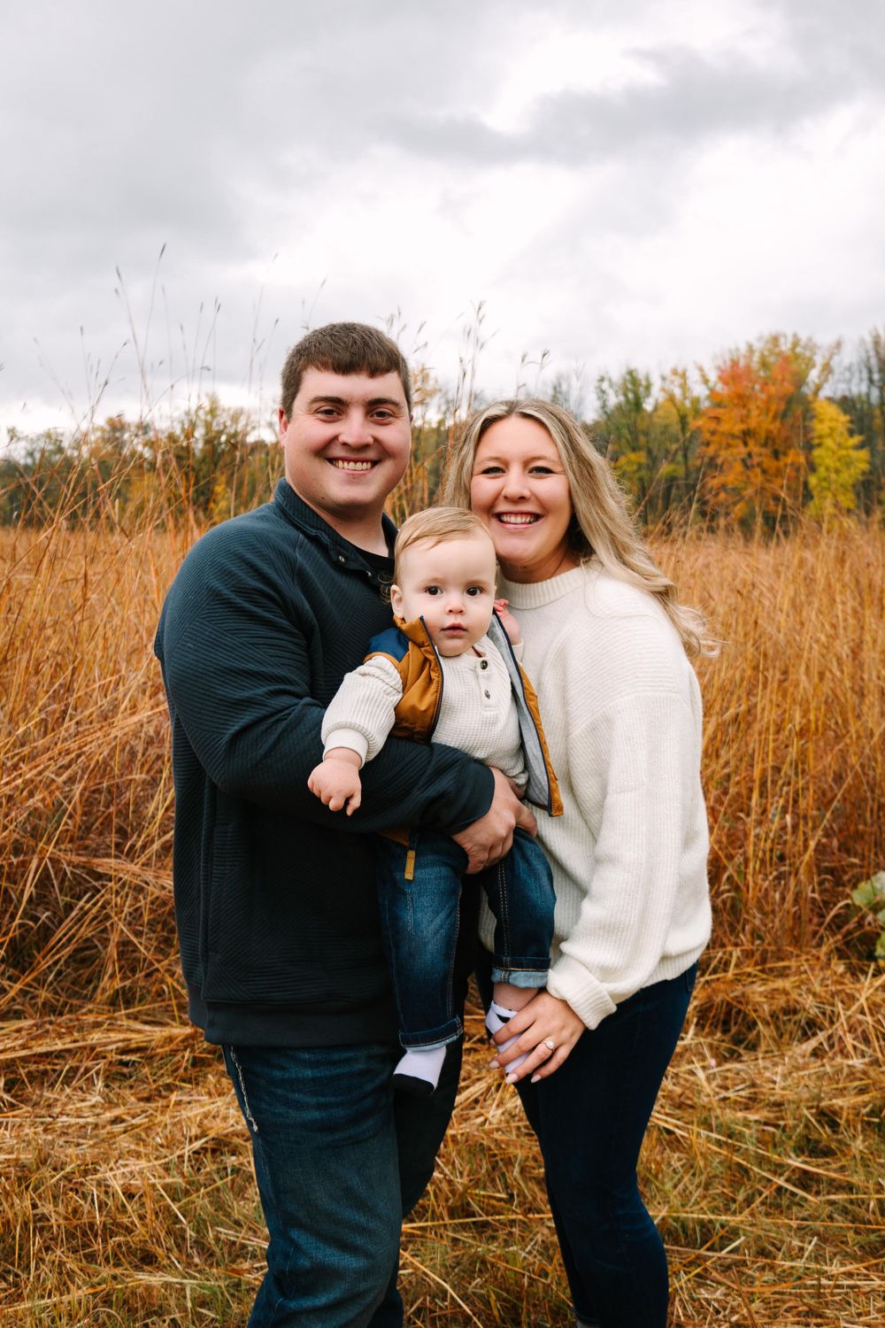 A family stands together in a field of tall autumn grass with fall foliage in the background.