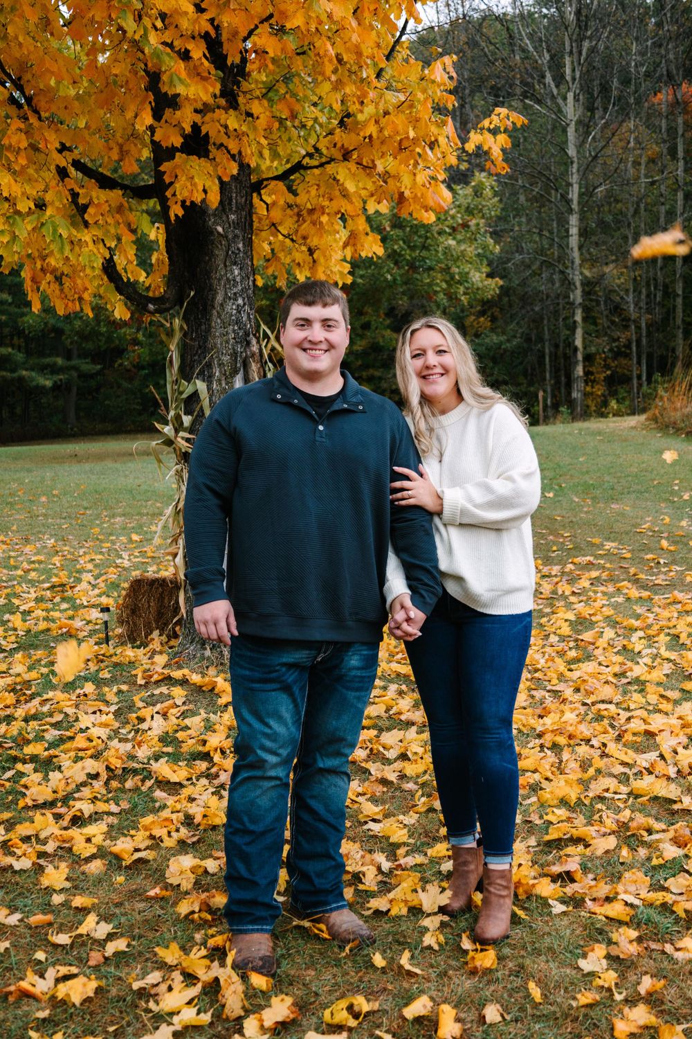 A couple stands together on autumn leaves beneath vibrant orange maple trees in a forest setting.
