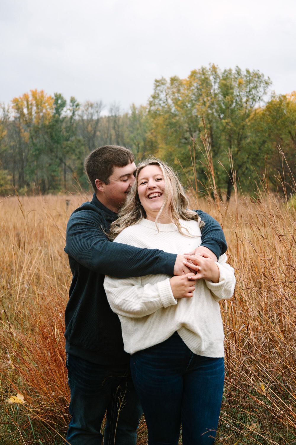 A couple embraces and laughs together in a fall field with tall golden grass and autumn trees in the background.