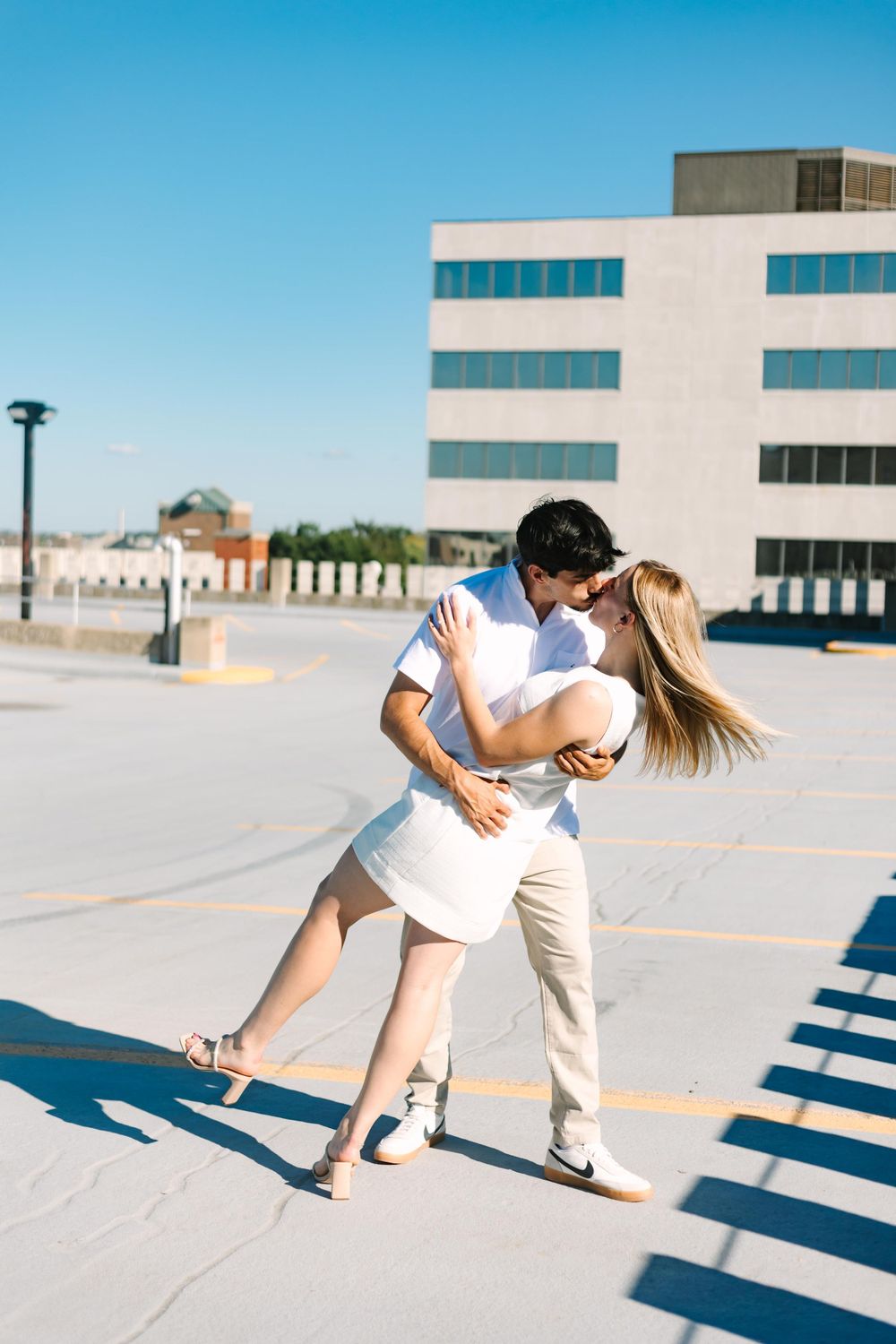 A romantic couple dances together on a rooftop parking lot with a modern building in the background on a sunny day.