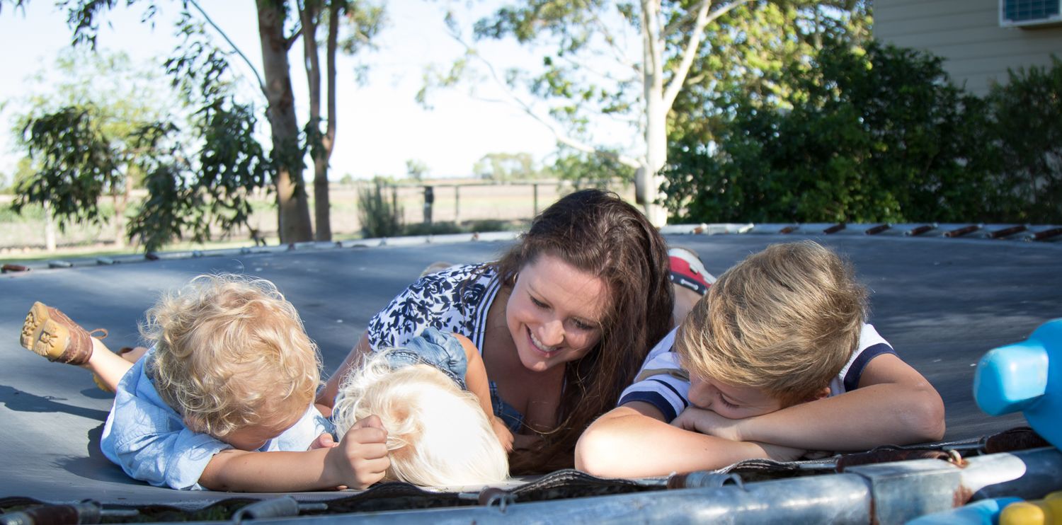 A woman and two children are lying on a trampoline, smiling and playing with a small dog. The background shows trees and a house.