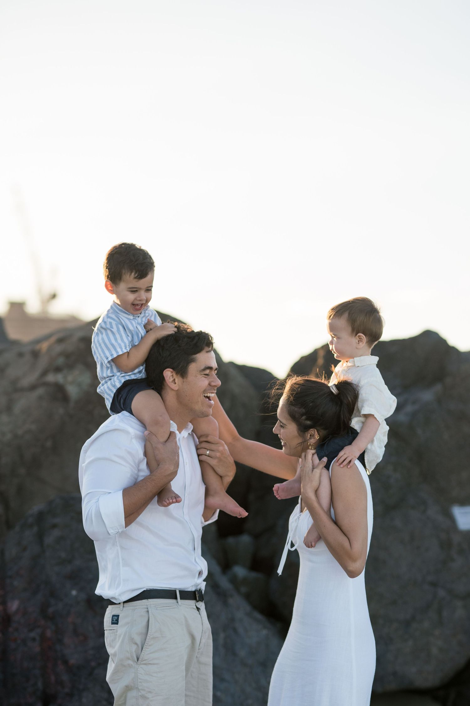 Parents laughing together on the beach, each carrying a little boy on their shoulders