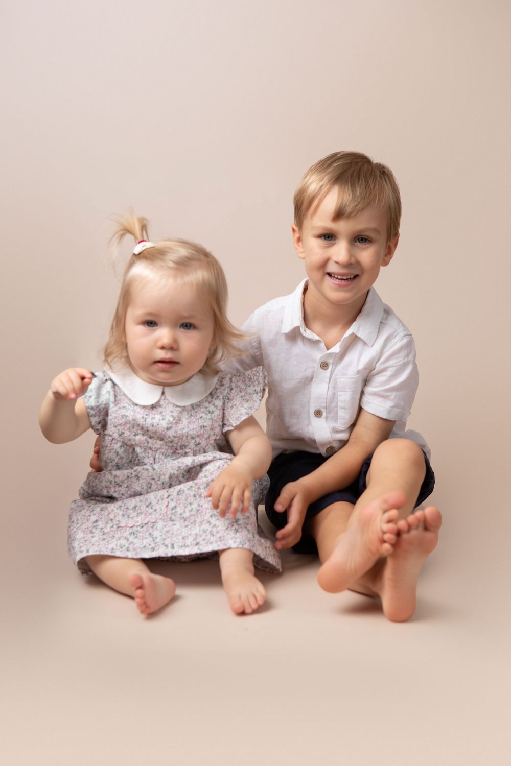 Adorable toddler girl in floral dress sitting with smiling older brother in studio portrait.