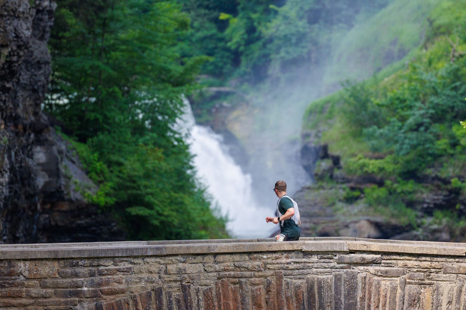 A person sits on a stone wall overlooking a misty waterfall surrounded by lush green forest.