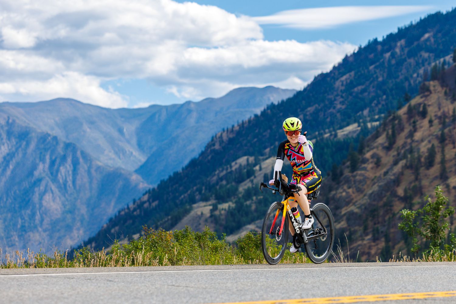 Mountain biker performs aerial jump trick on dirt trail with scenic mountain range backdrop in clear blue sky.