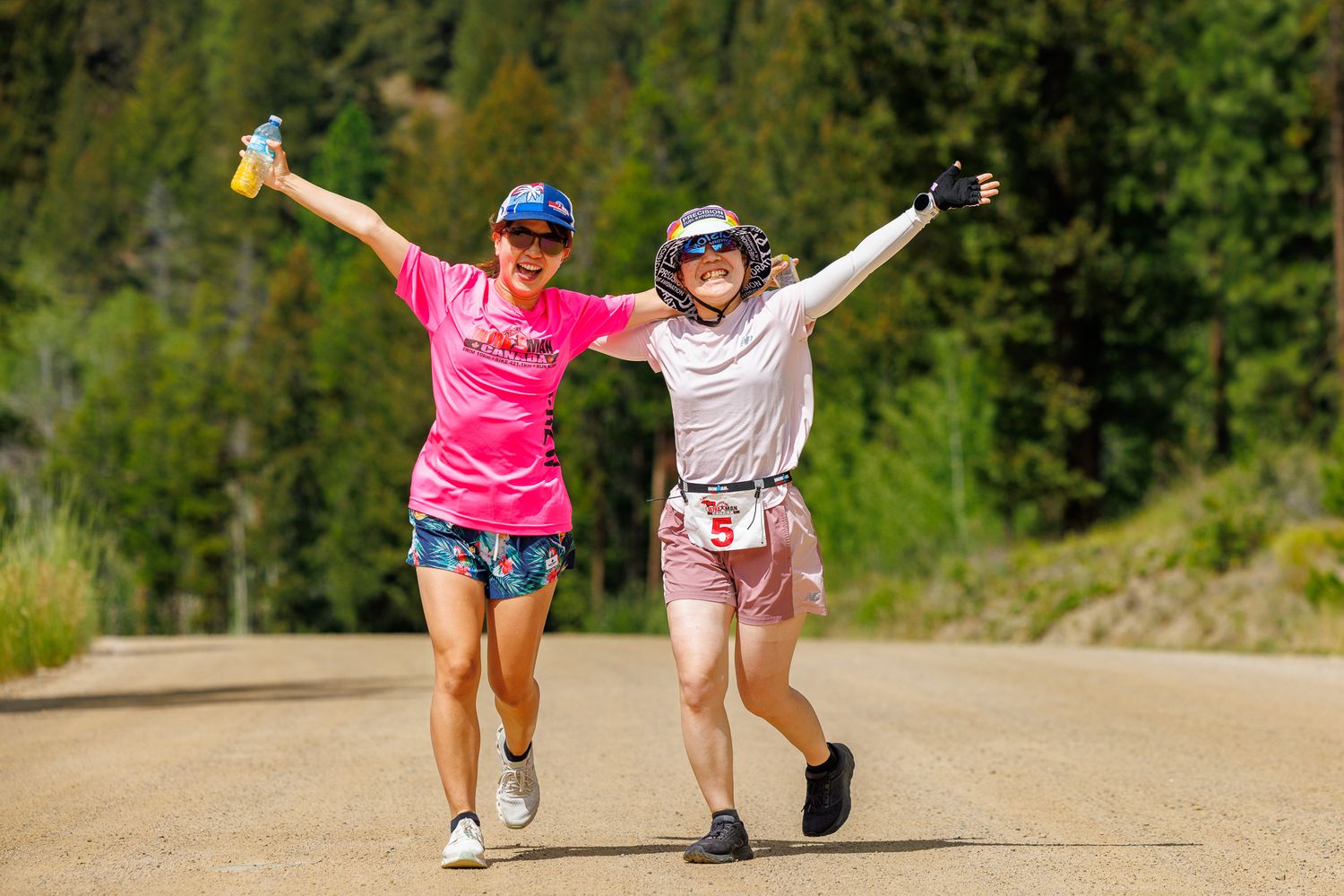 Two people in athletic wear run joyfully down a dirt trail surrounded by lush green trees and vegetation.