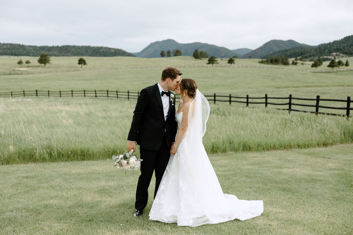 Romantic wedding couple embraces on rolling Colorado hills with mountains and rustic fencing in the background.