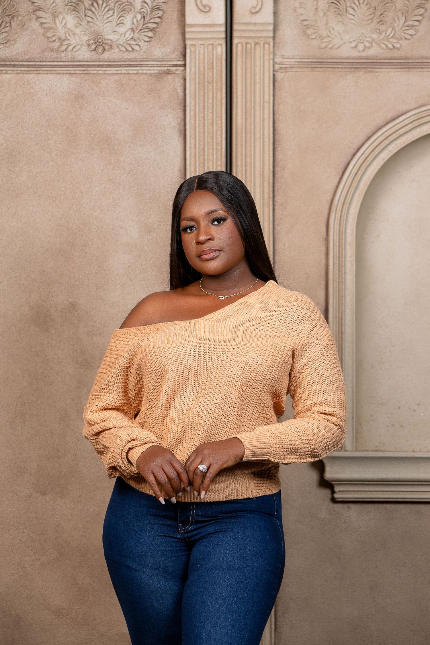 Casual portrait against ornate wall wearing peach off-shoulder sweater and blue jeans.