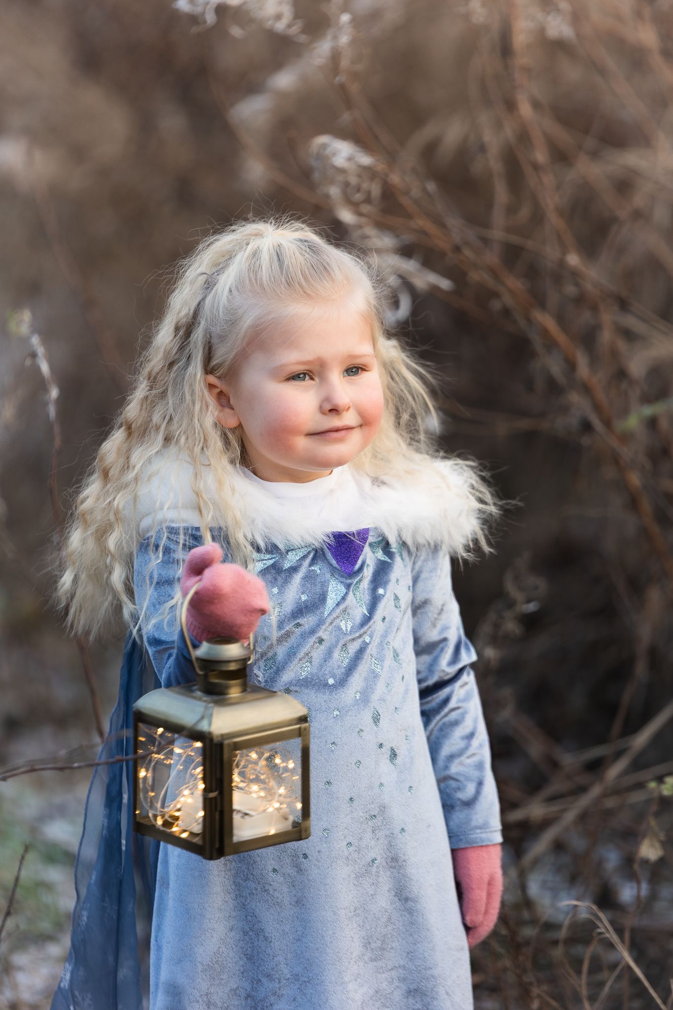 Child in light blue winter dress holding glowing lantern against snowy background.