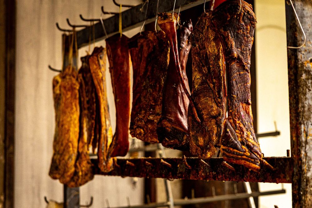 Rows of smoked and cured meat cuts hanging on metal hooks in a rustic smokehouse.