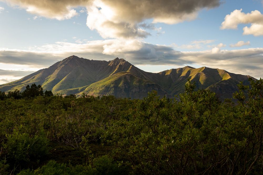 Sunlit green mountain range with dramatic clouds casting shadows over lush tropical vegetation.