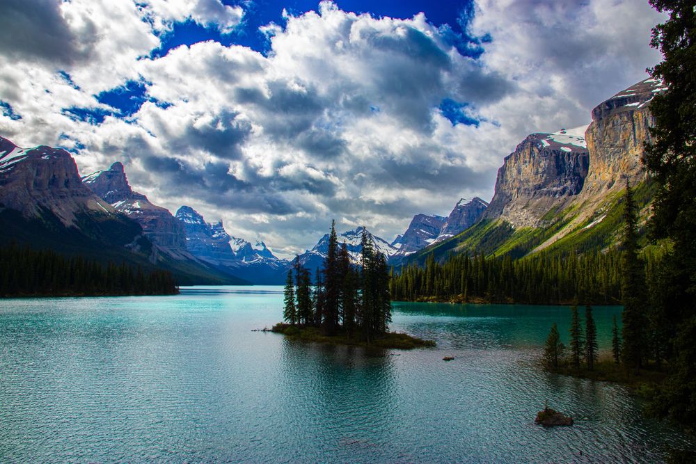 Turquoise glacial lake with a small pine-covered island surrounded by towering Rocky Mountains.