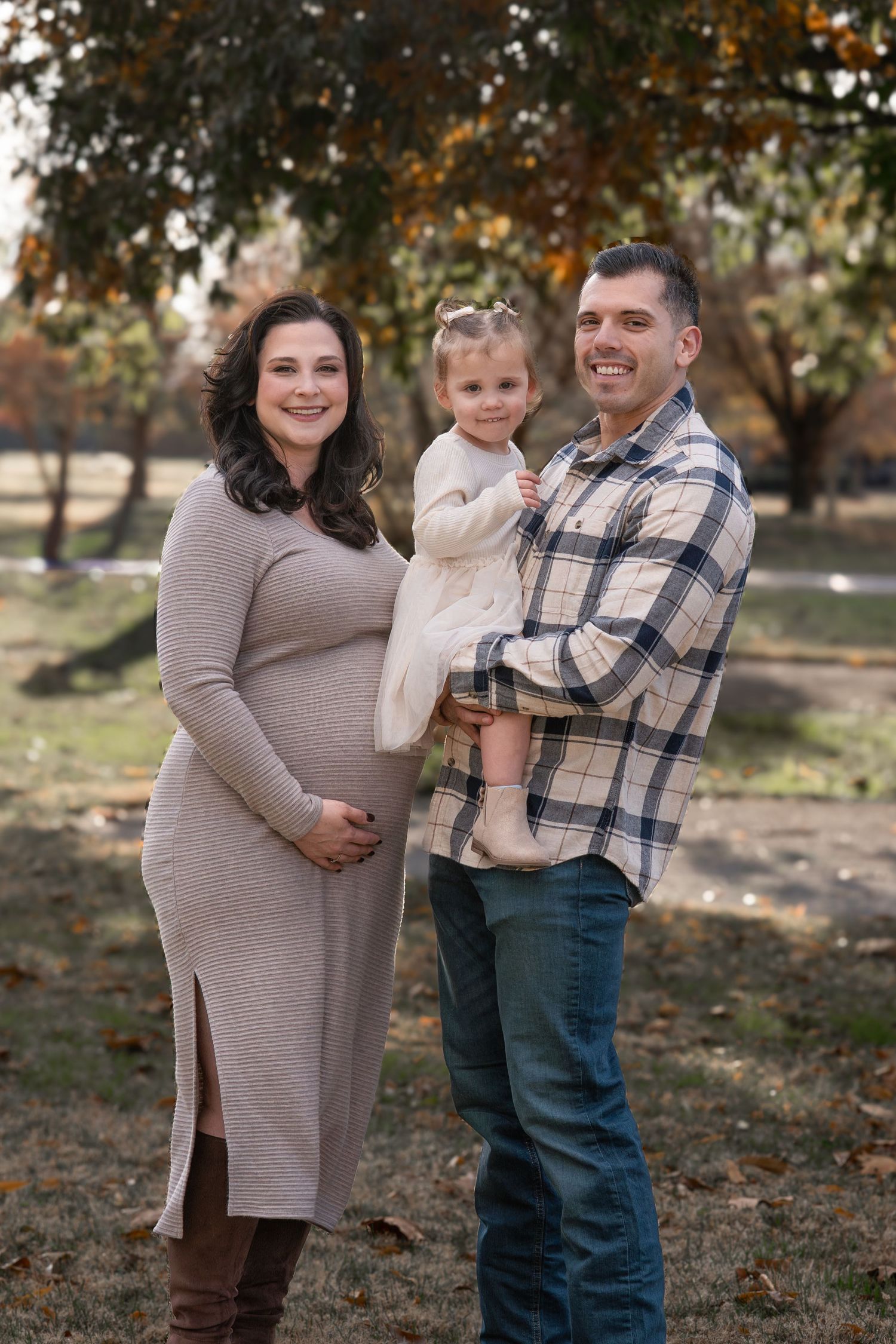 A family poses together outdoors during autumn, with a pregnant woman in a sweater dress and a toddler held by her father.