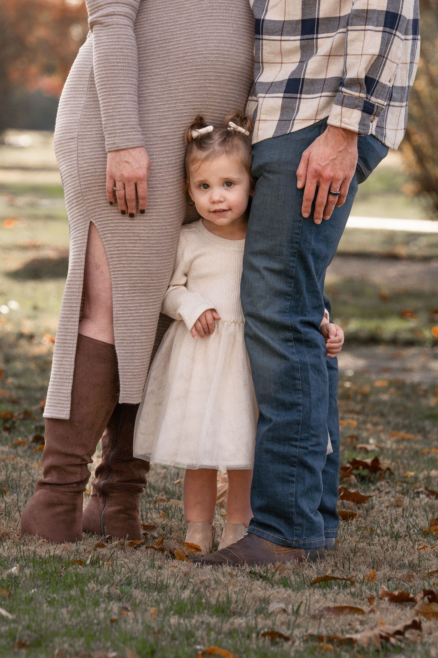 A family's feet and legs stand together on fallen autumn leaves, with a little white dress visible.