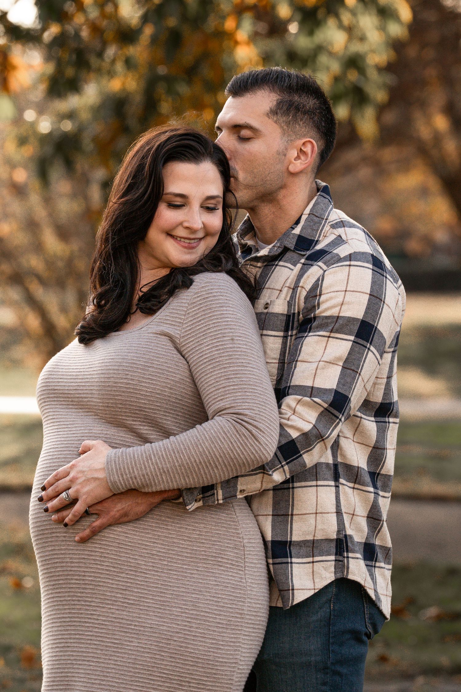 A couple embraces outdoors during an autumn maternity photoshoot wearing a striped sweater and plaid shirt.