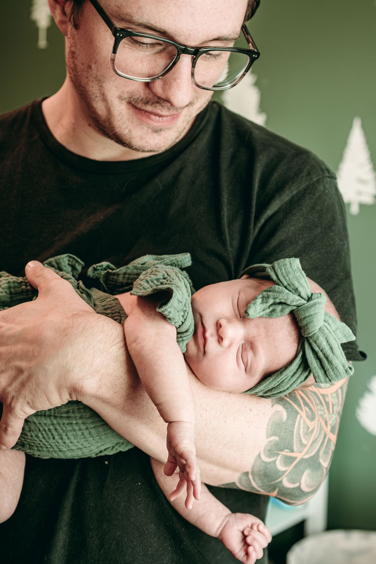 A person in a black shirt holds a sleeping newborn baby wrapped in a green blanket with a matching bow headband.
