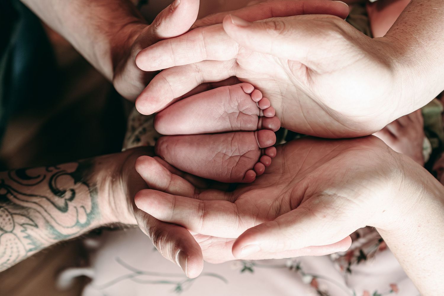 Close-up of tattooed hands gently holding tiny newborn feet against soft fabric background.