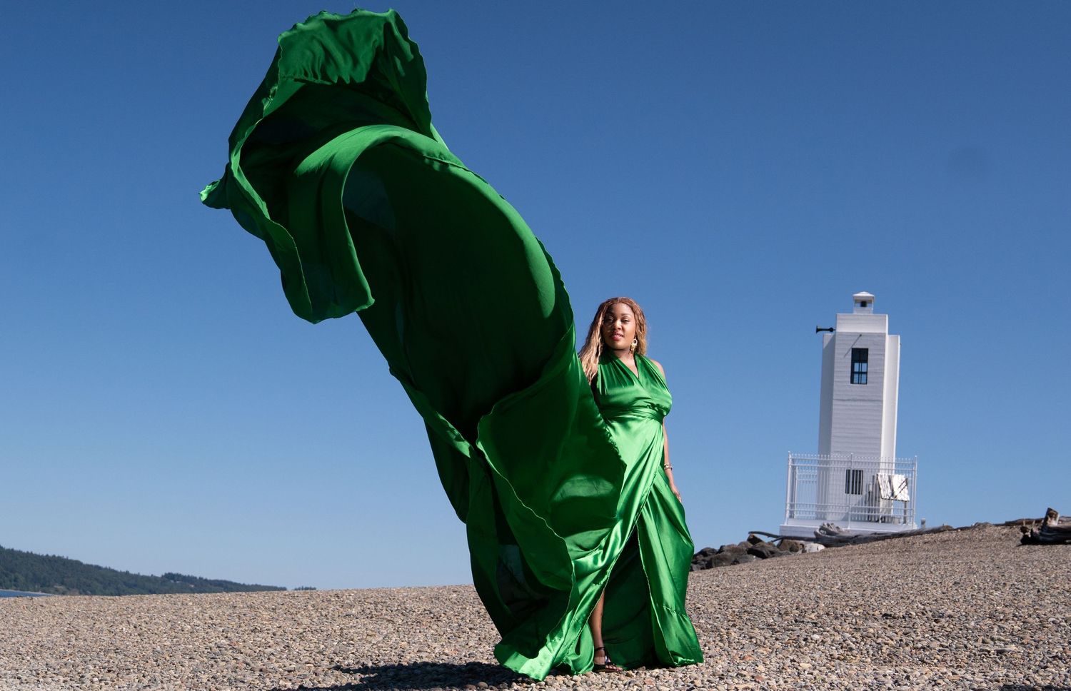 Flowing emerald green dress billows dramatically against blue sky and lighthouse backdrop.