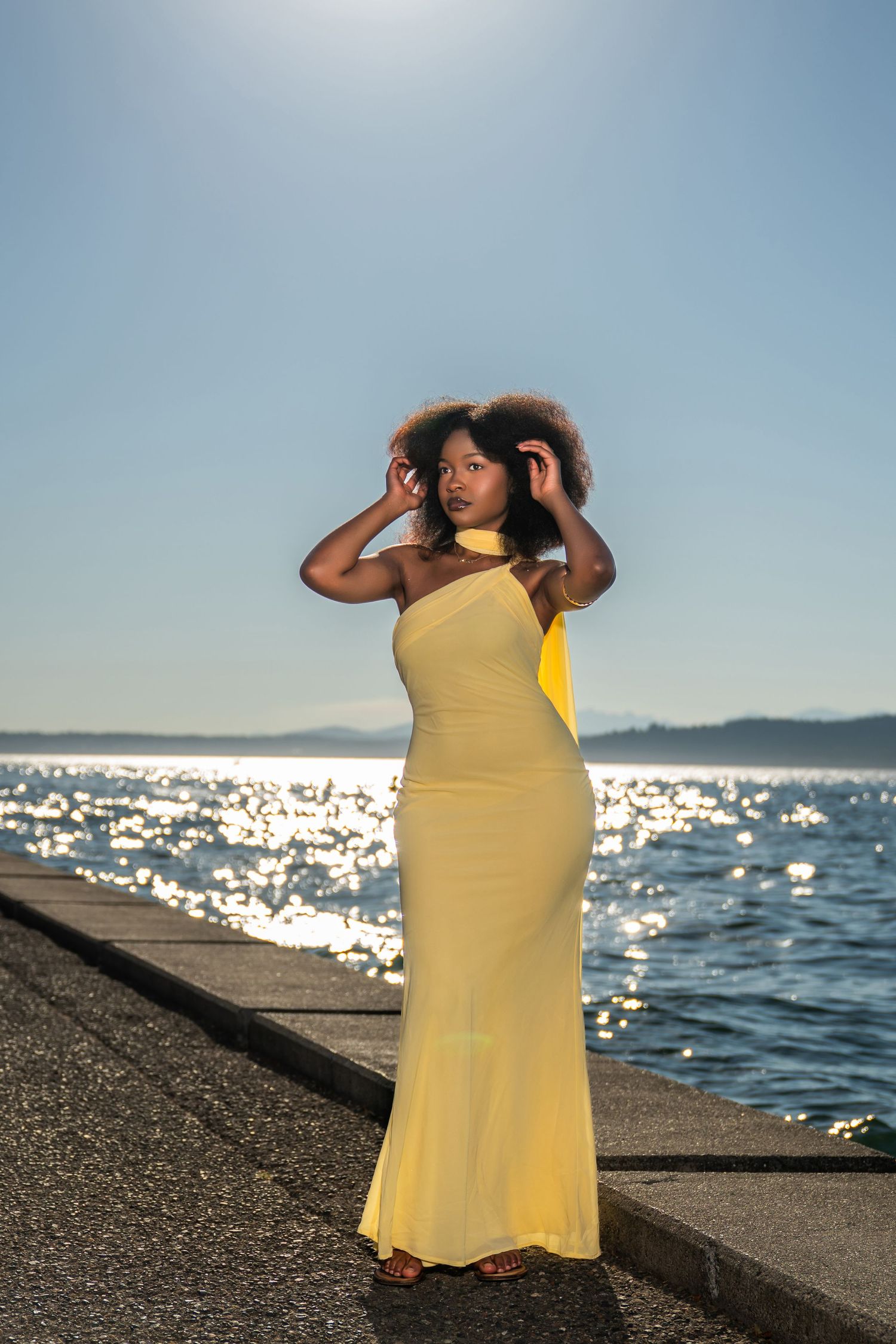 A person in a yellow dress stands on a beach seawall looking out at the sparkling ocean waters at sunset.
