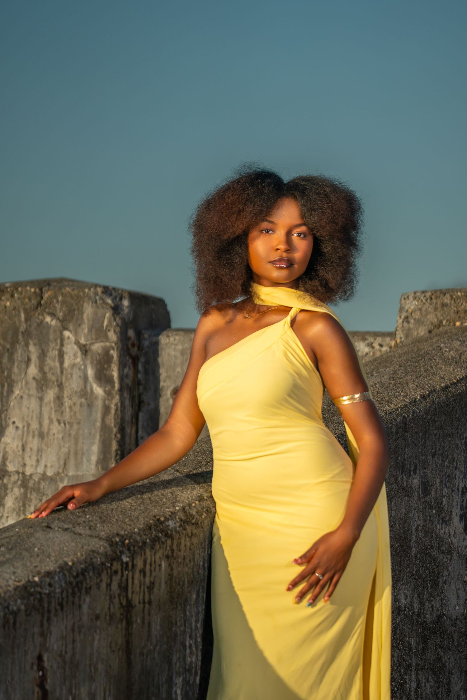 A figure in a flowing yellow dress poses dramatically against a stone wall at sunset.