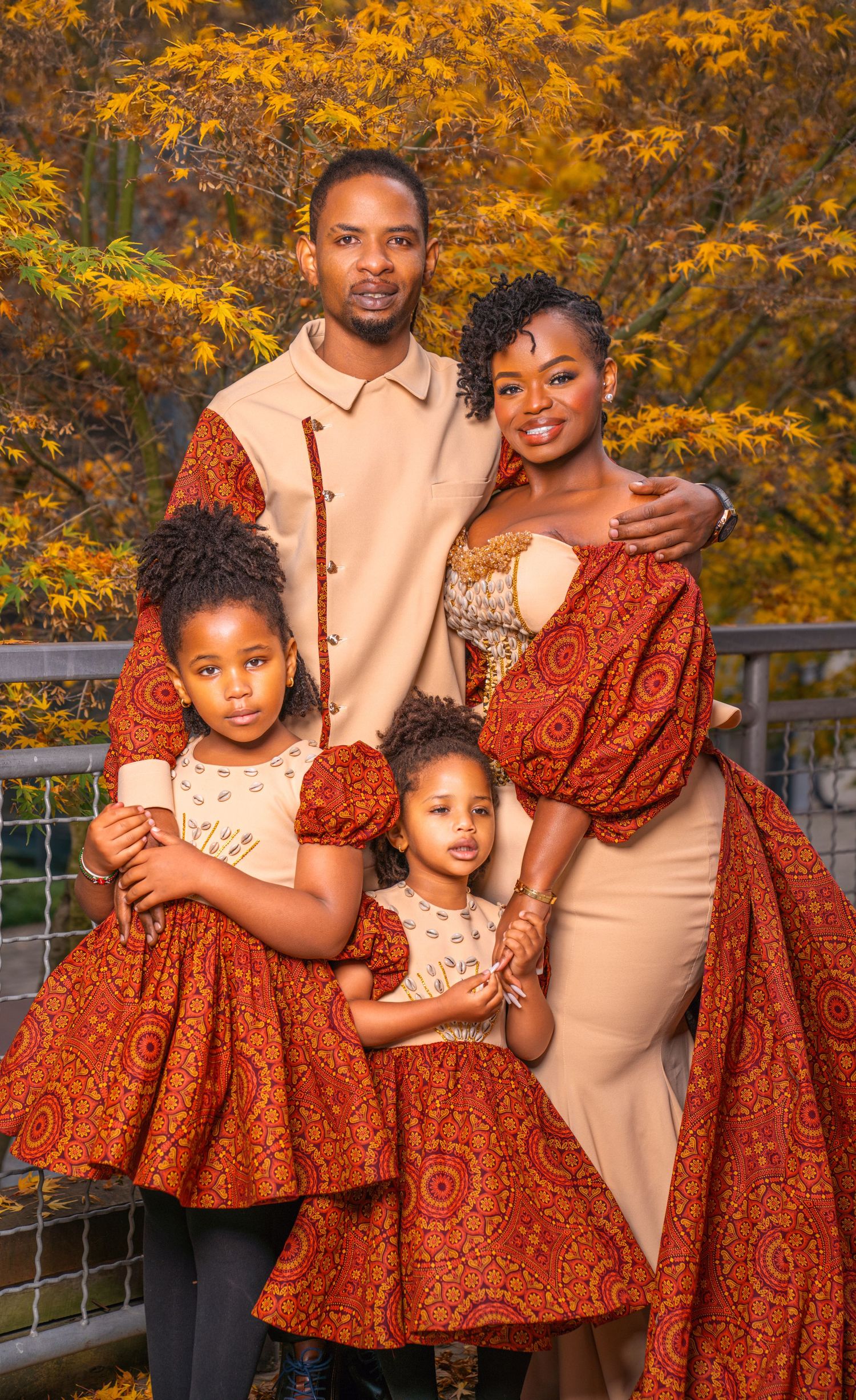 Family wearing coordinated orange and beige outfits poses together against autumn foliage backdrop during sunset.