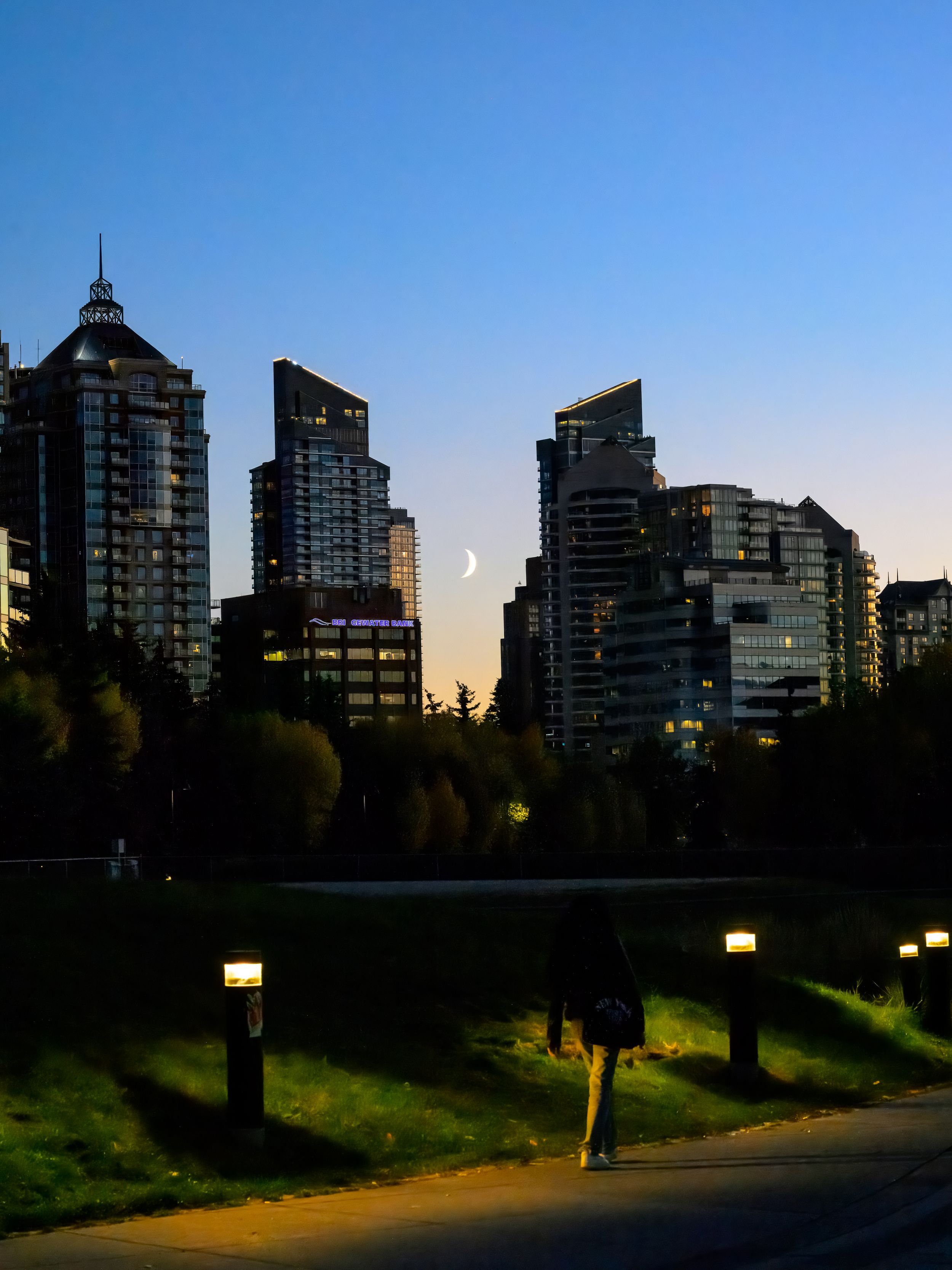 Urban skyline silhouette at dusk with illuminated high-rise buildings and walking path along a park.
