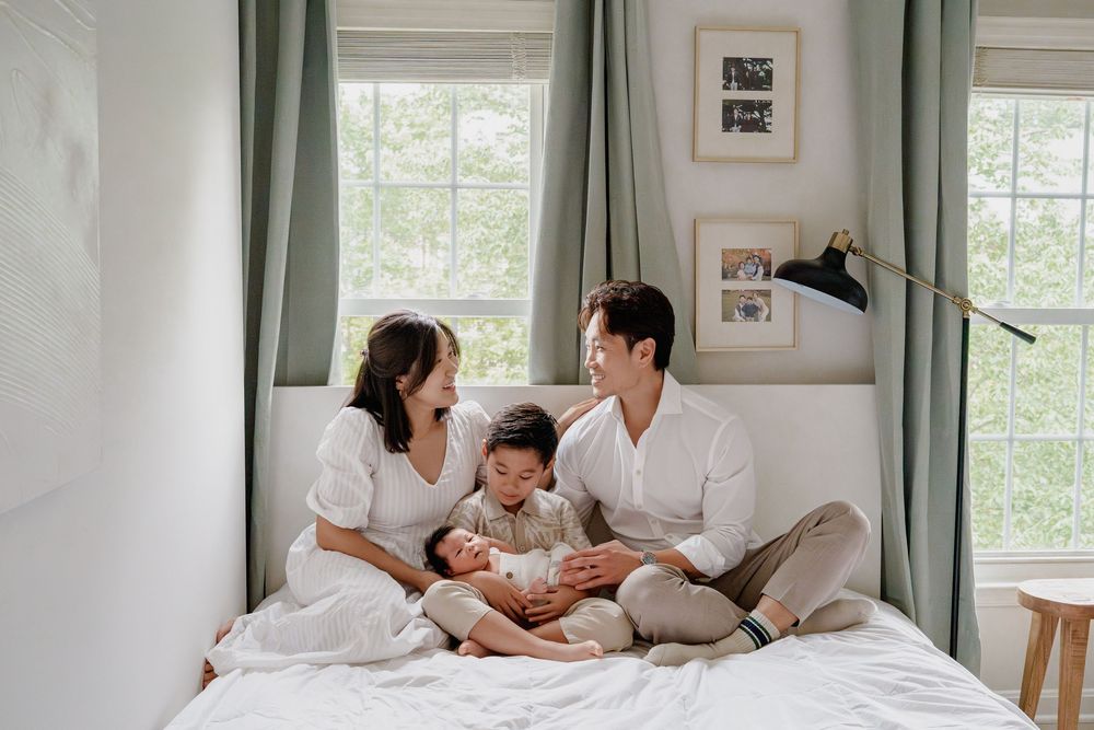 Happy Asian family of four sitting on a white bed near windows, parents with toddler and newborn baby, in Fairfax, VA