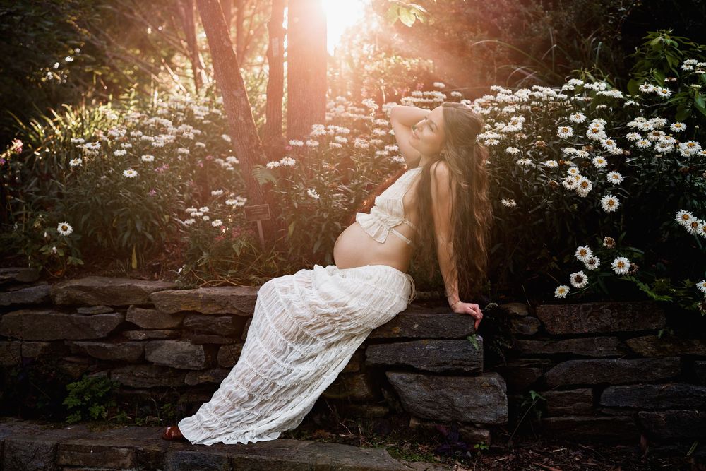 Pregnant woman in white dress sits on stone wall surrounded by wildflowers and daisies in golden sunlight, in Maryland