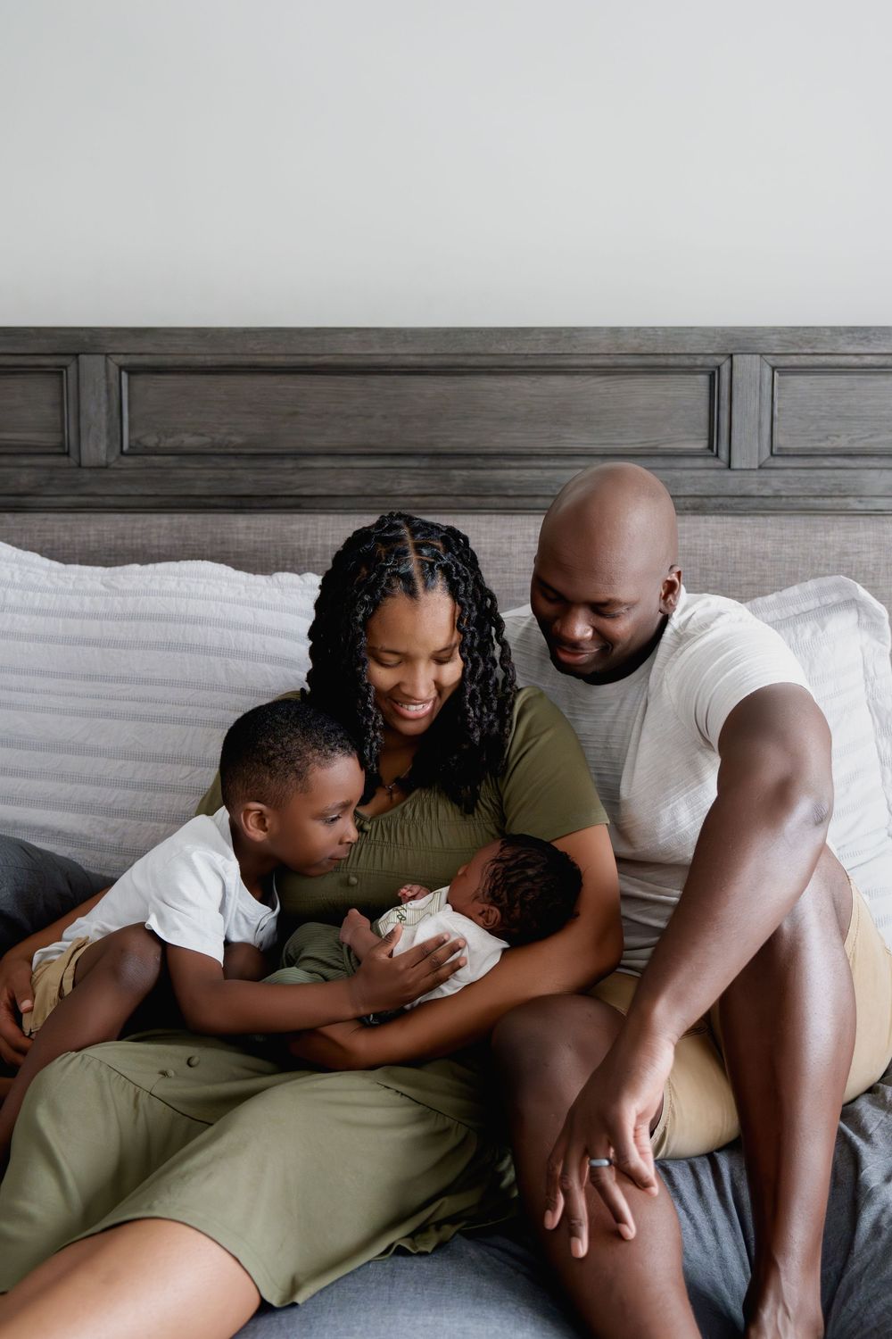 Happy Black family sitting on bed, parents smiling at newborn baby while young son looks on lovingly, in Hanover, MD