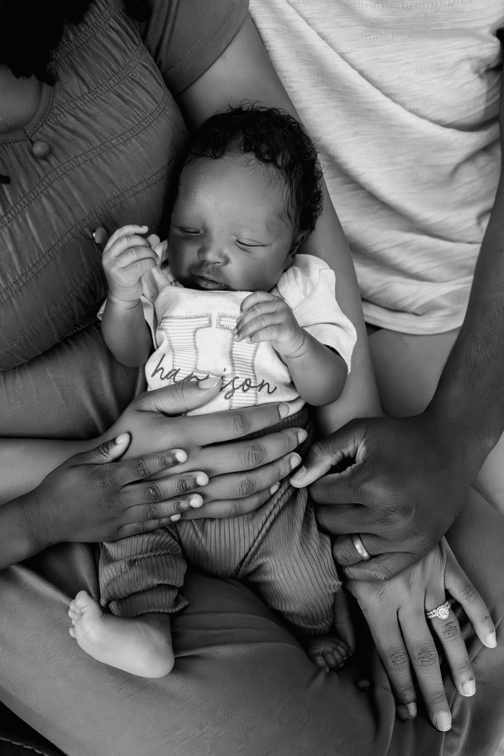 Black and white photo of a sleeping newborn baby named Harrison cradled in parents hands wearing a personalized onesie, in Hanover, MD