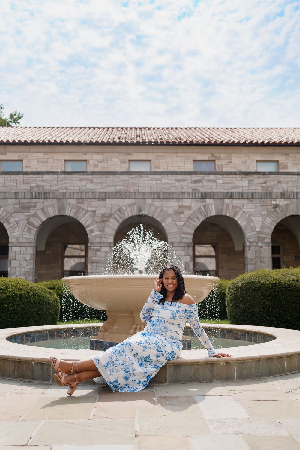 Pregnant woman in blue floral dress sitting by ornate stone fountain in elegant courtyard with arched architecture, in Ellicott City, MD