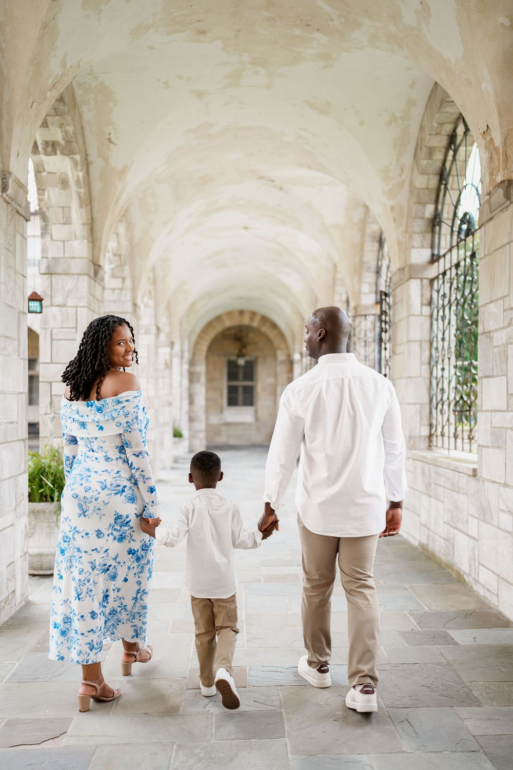A family of three walks through an elegant stone archway corridor, holding hands, mother in blue floral dress, in Ellicott City, MD