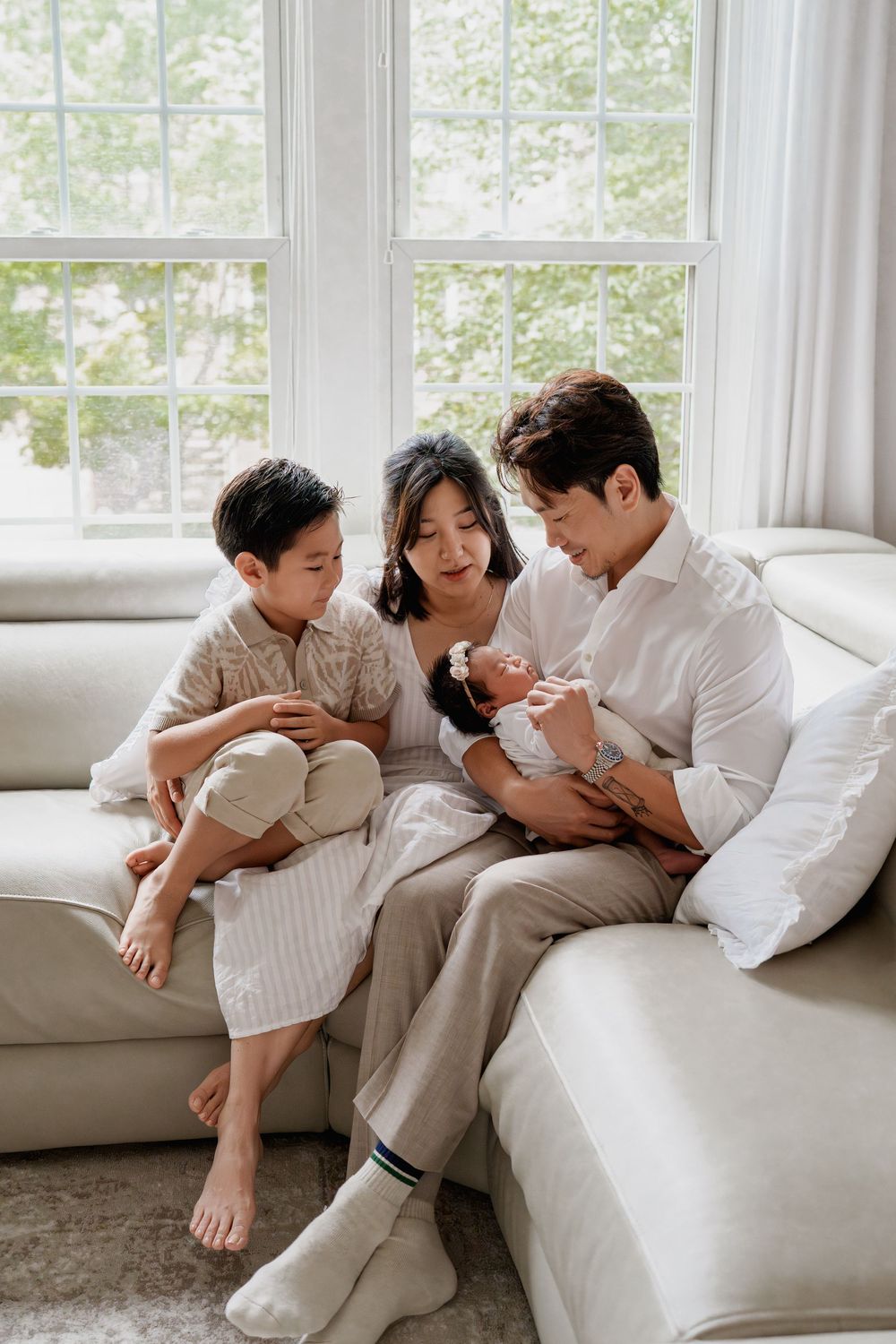 Family of four sitting on a white sofa, parents gazing at their newborn baby while their young son looks on warmly, in Fairfax, VA