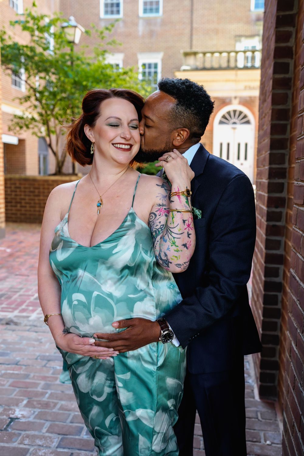 A man in a navy suit kisses a smiling pregnant woman in a floral green dress on a brick-lined street, in Alexandria, VA