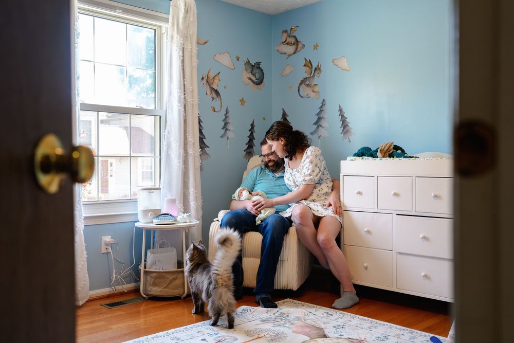 Parents sitting in nursery chair with newborn baby, dragon wall decals on blue walls, white dresser, and small dog nearby, in Baltimore, MD