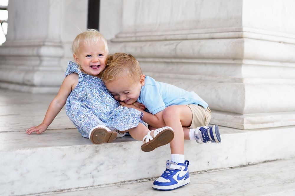 Two toddlers in blue outfits sitting on marble steps, sharing a sweet hug outdoors, at the Jefferson Memorial in Washington DC