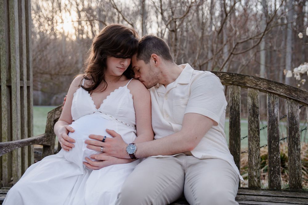 Couple in white outfits sharing tender moment on bench, hands on pregnant belly, surrounded by trees at sunset, in Wheaton, MD