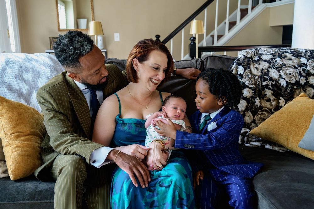 Family of four sitting on sofa, parents smiling at newborn baby while young child in suit looks on, in Silver Spring, Maryland